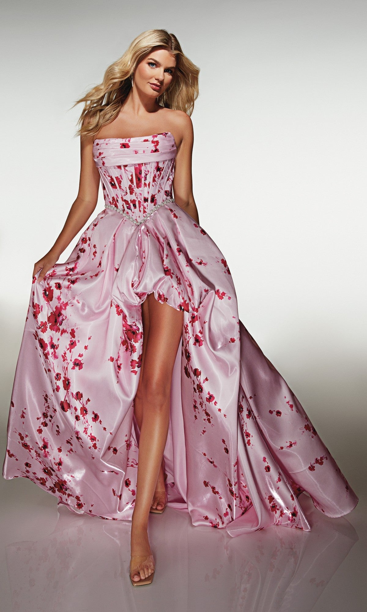 A woman wears the Alyce 61989 long prom dress—strapless, pale pink satin with red floral patterns, a corset bodice, and high-leg slit—while standing against a neutral background.